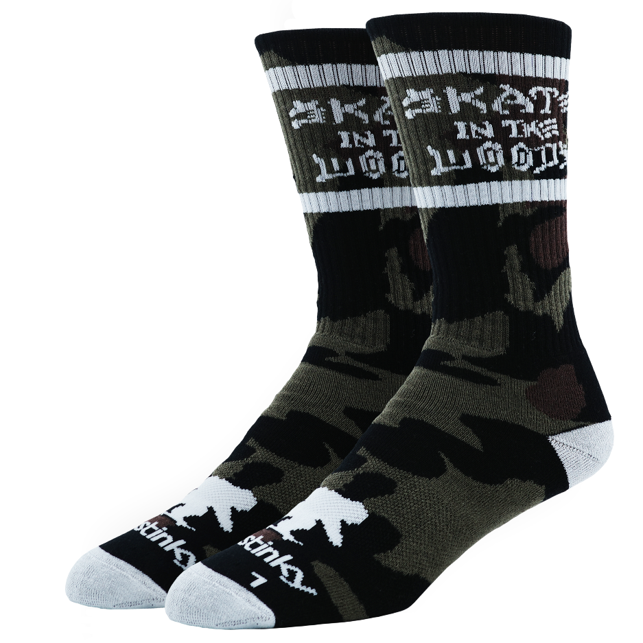 A pair of crew socks featuring a black and olive green camouflage pattern, with white text reading 'SKATE IN THE WOODS' on the upper cuff and white accents on the toes and heels, set against a plain white background.