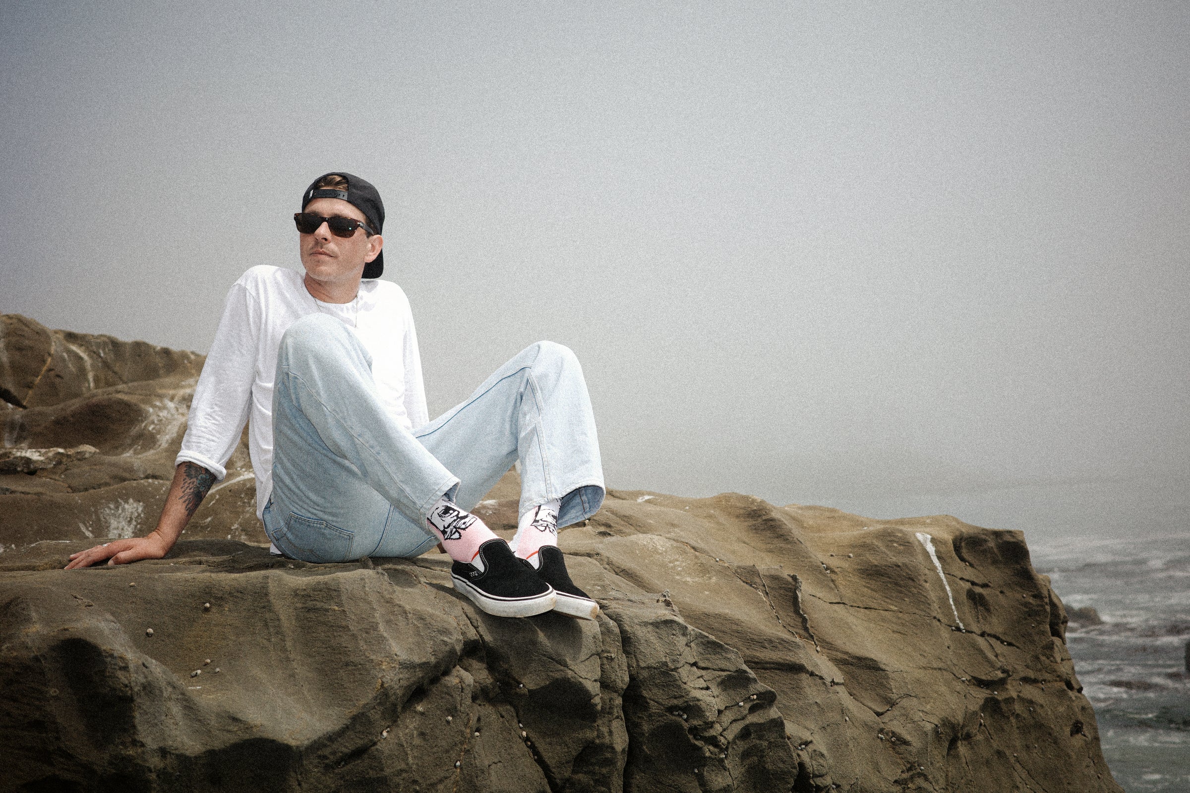 Person sitting on a rocky outcrop by the ocean wearing sunglasses and casual clothing. With Stinky Socks 