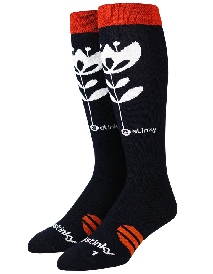 A pair of black knee high socks with a white stylized flower design, orange cuffs and heel accents, and white/orange 'jstinky' branding on a plain white background. Made out of light merino wool fibers